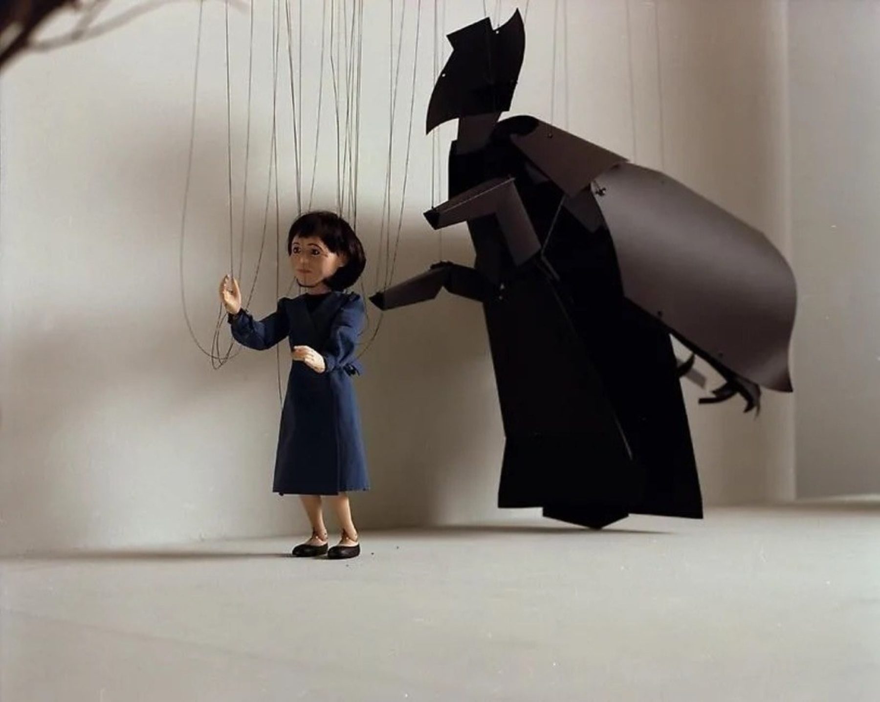 A still from a puppet show of a woman in a blue dress with short dark hair being followed by a black ghost-like creature.